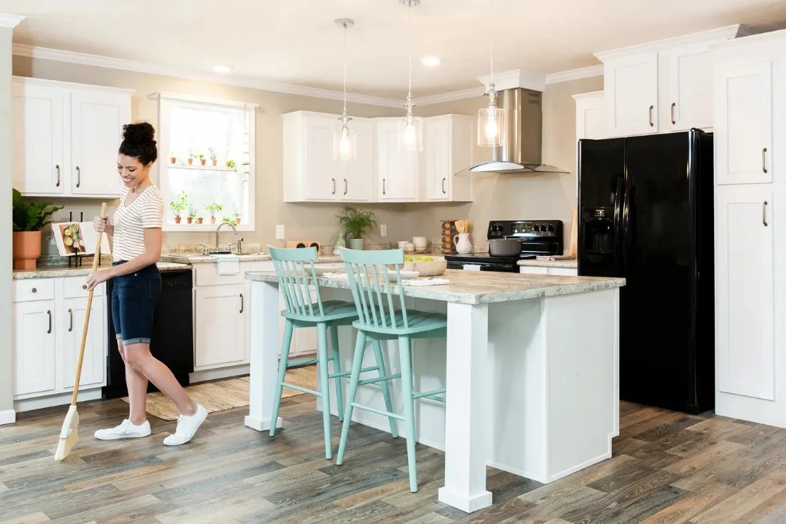 A woman sweeps the floor in a modern manufactured home kitchen with white cabinets and a kitchen island.