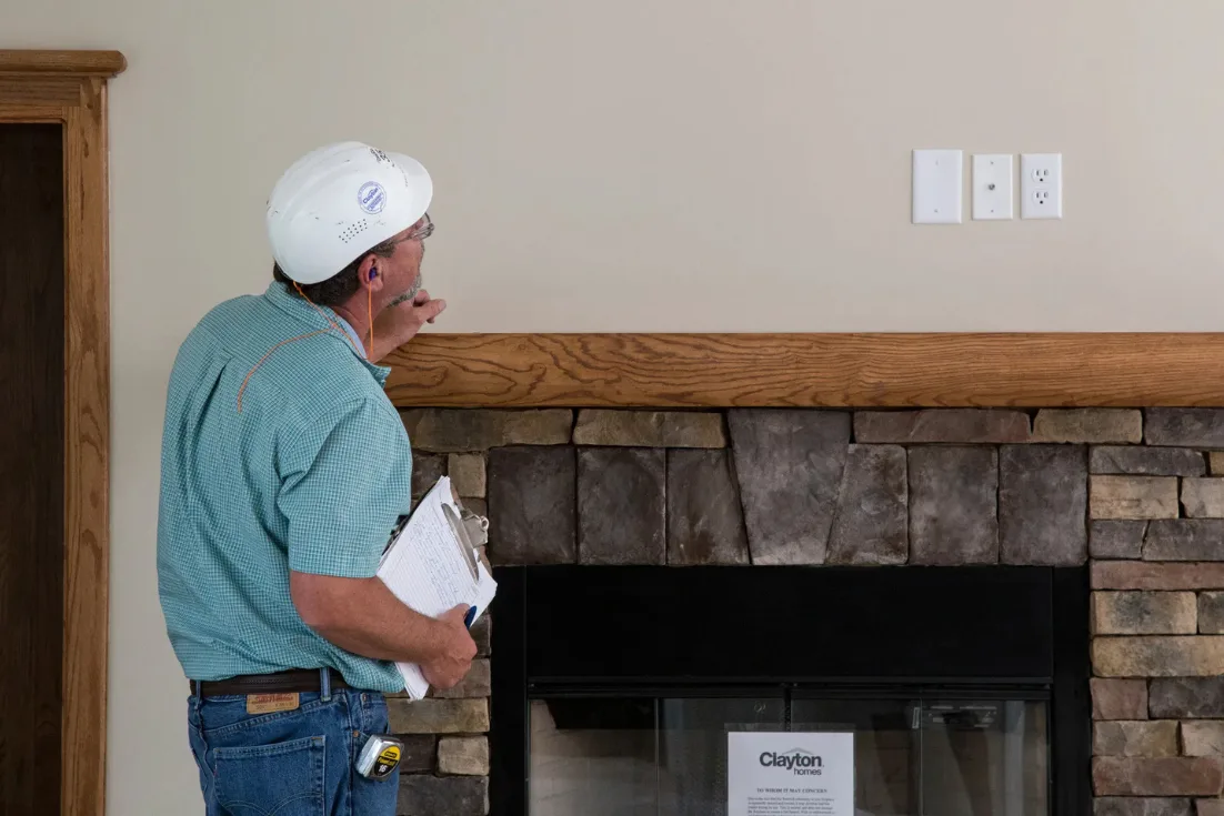 An inspector in a blue shirt and white construction hat holds a clipboard and looks at the wall above the mantle piece of a stone fireplace.