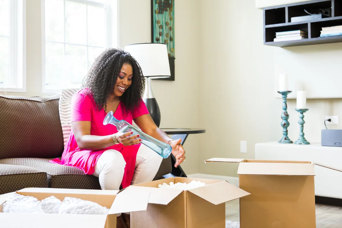 A smiling woman in a pink blouse sits on a couch wile packing a glass vase one of the moving boxes in front of her.