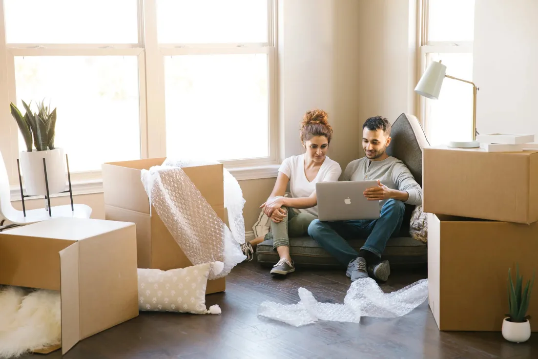 A couple sits inside looking at a laptop with packing boxes around them as they prepare to move into their new home.