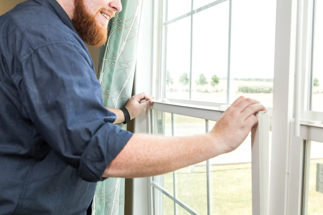 A man seen from his red beard down opens a grid-paned window from inside a home.