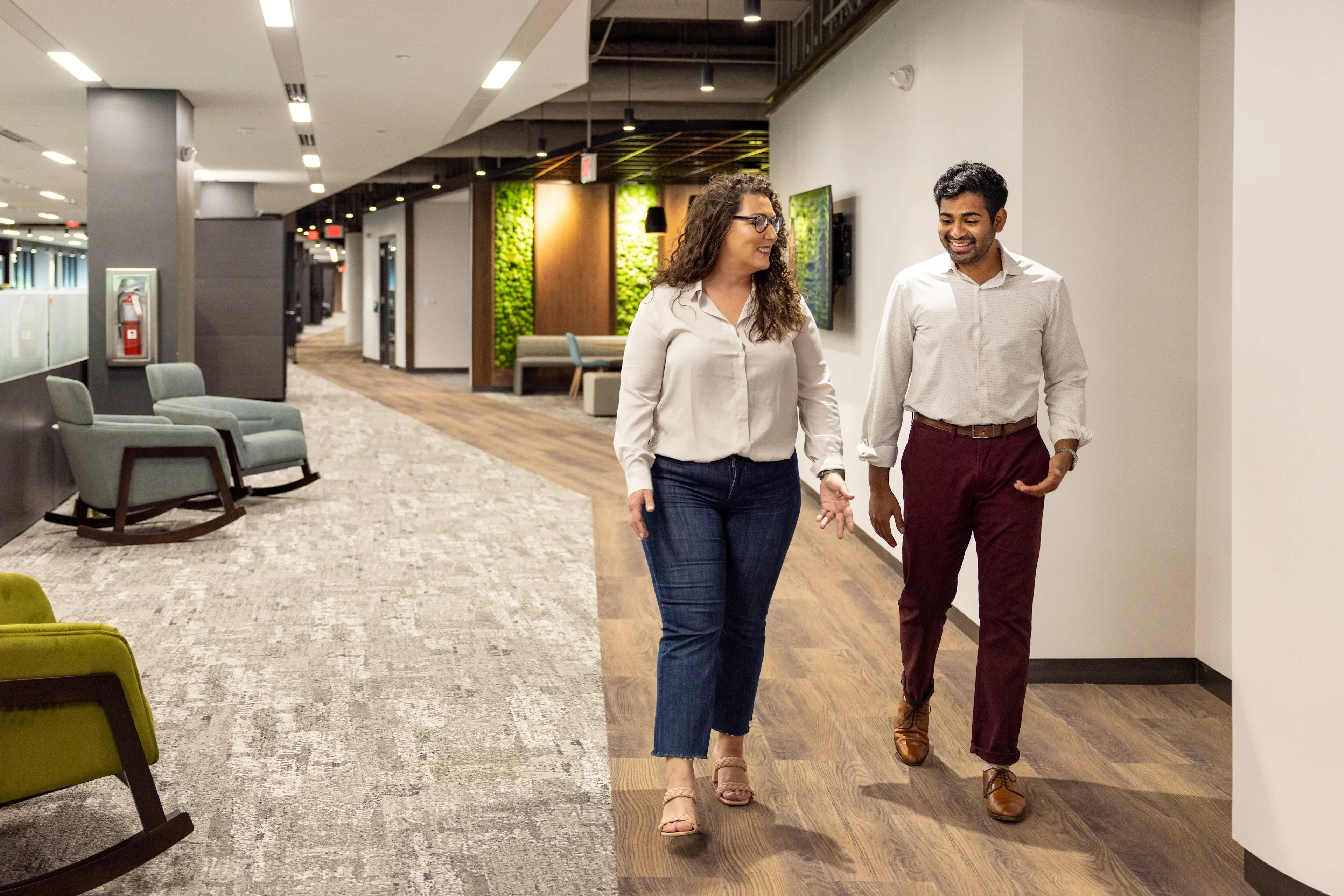 Man and woman in white shirt and pants wall down the hallway of an office with chairs and a plant wall in the background.