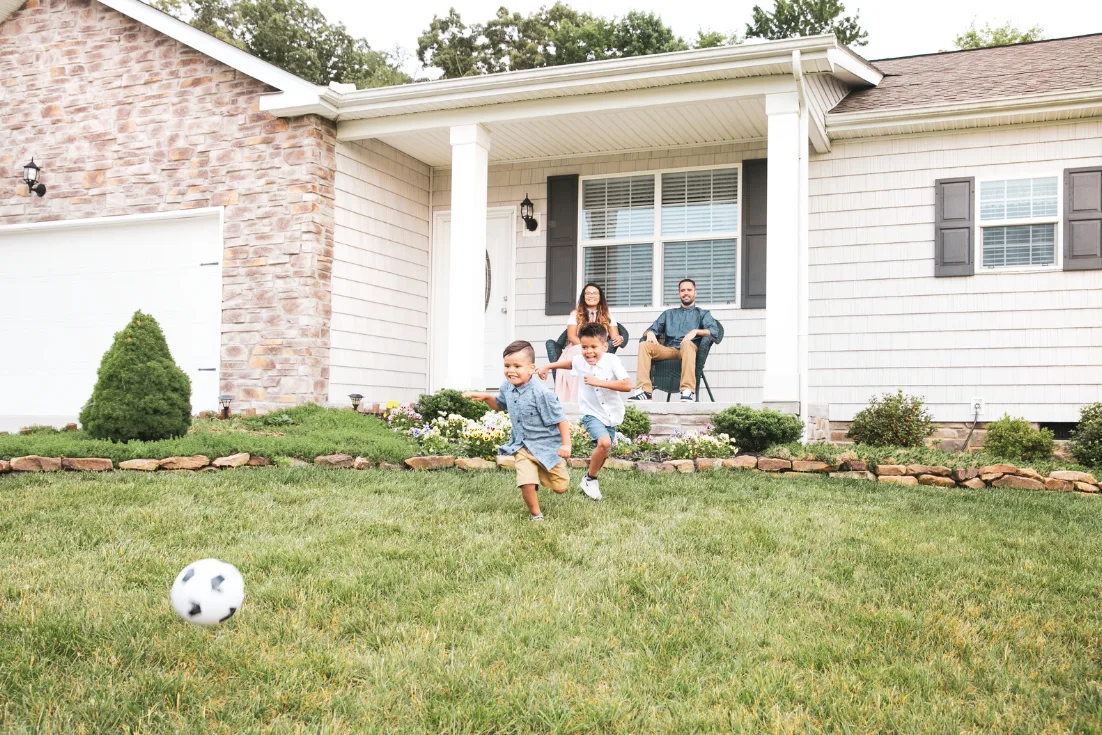 Two young boys run after a soccer ball in a front yard while their parents sit in chairs on the front porch of a home with beige siding and a brick garage.
