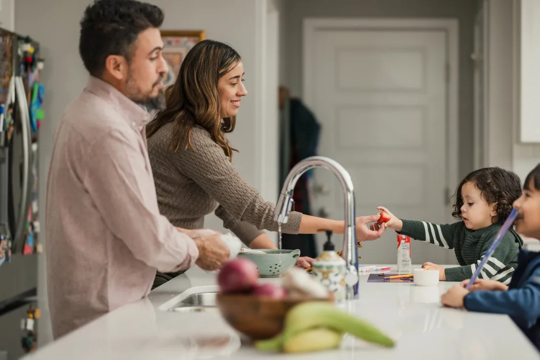 A dad washes dishes while the mom beside him hands over a strawberry to her toddler son over a kitchen island with marble countertop.