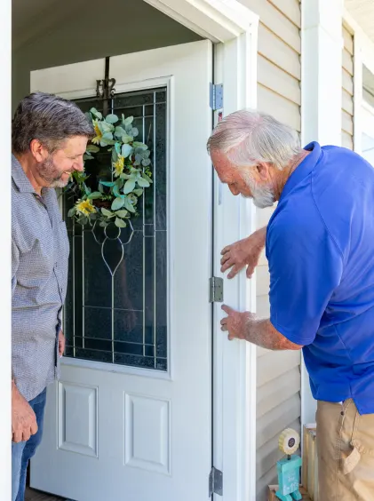 A homeowner stands in his open front doorway watching while a service person in a blue polo checks on the doorframe.