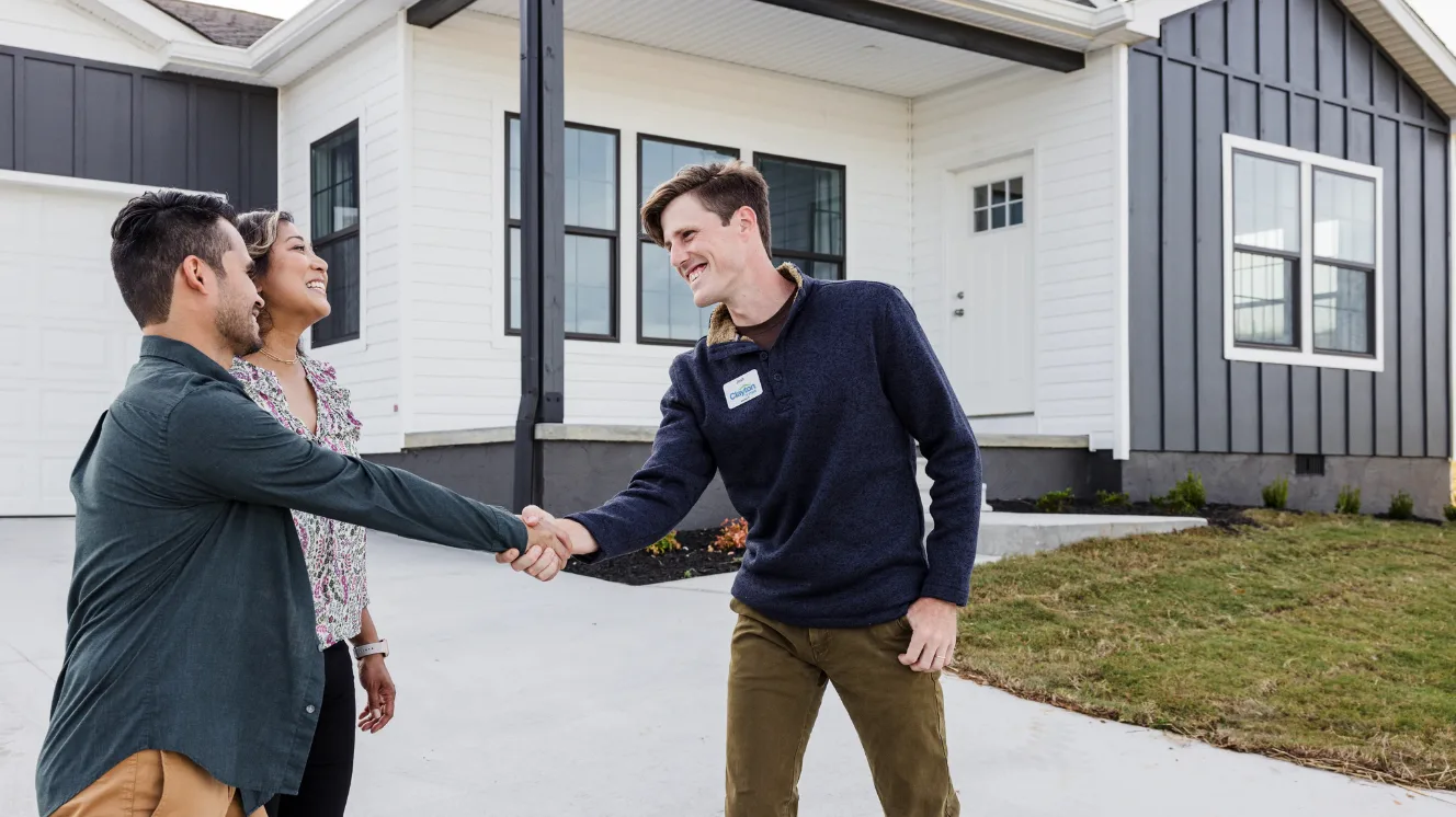 A couple shakes hands with a home consultant in front of a white and gray home with a front porch.
