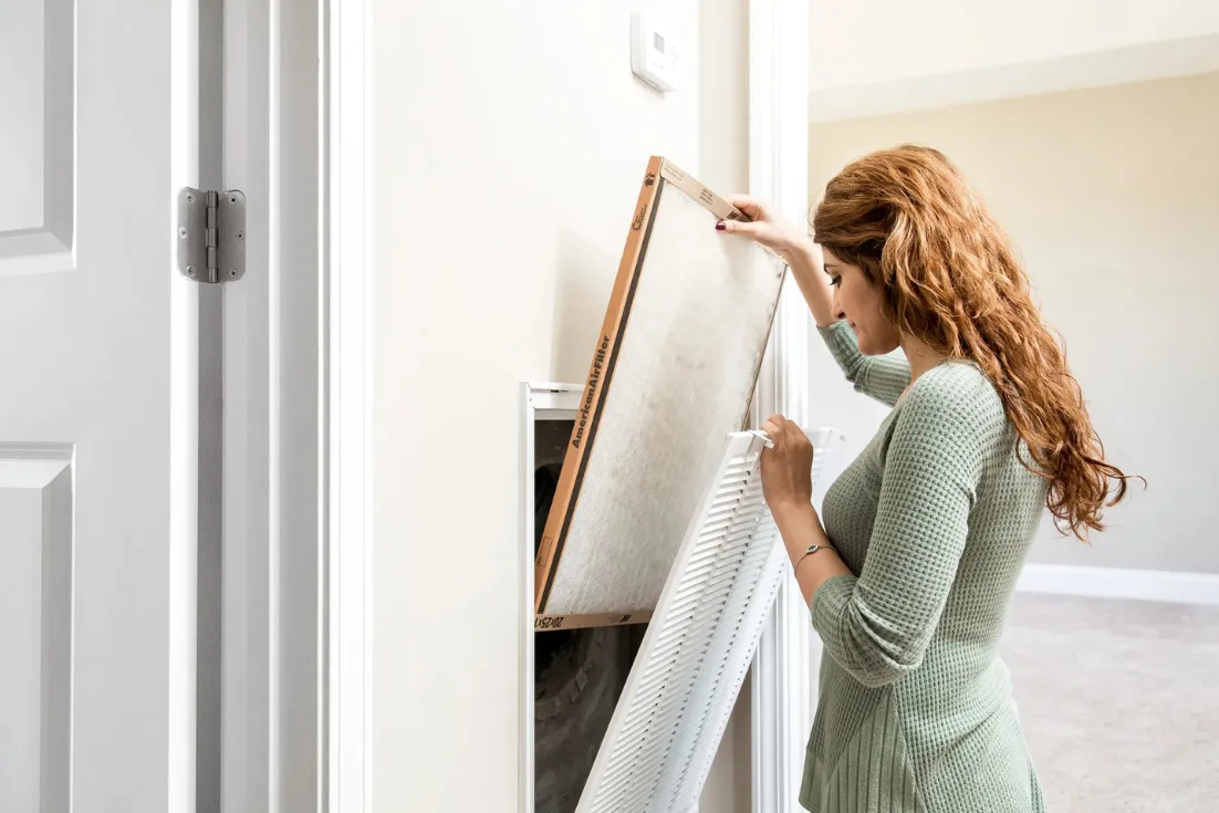 A woman in a light green sweater holds the filter grill with one hand while sliding a new air filter into place.