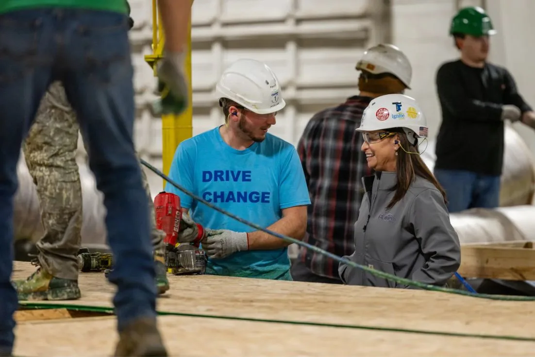 Lisa smiles while talking to facility worker in a blue shirt, both wearing white hard hats.