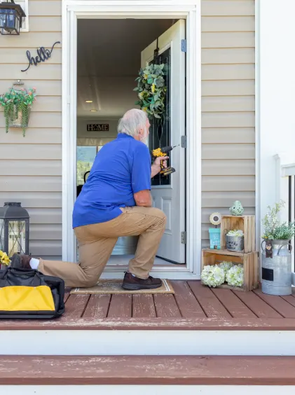 A service person in a blue polo kneels on one knee at an open doorway with a drill in one hand and a yellow tool bag beside him on the ground.