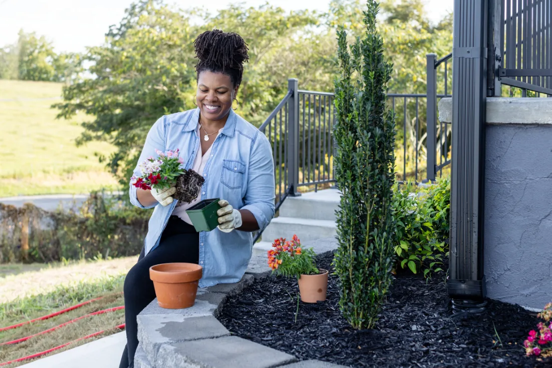 A woman in a chambray shirt pots a flowered plant beside her mulch bed.