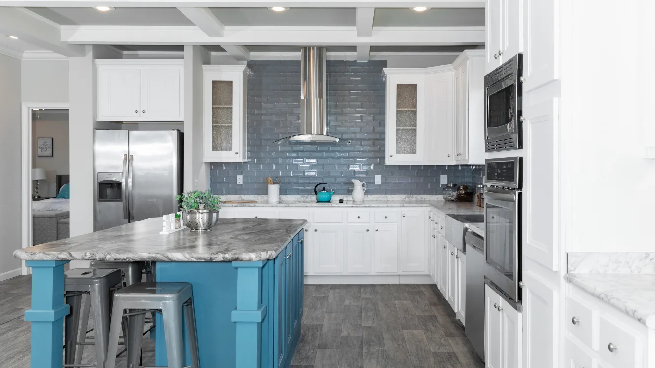 The kitchen of a manufactured home with a large blue island, stainless steel appliances, white cabinets, gray backsplash, and quartz countertops.
