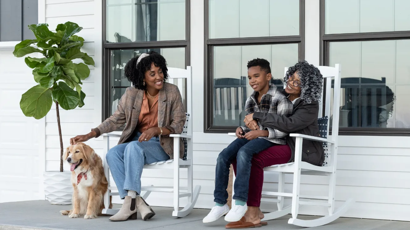 A family sits in white rocking chairs on a home's front open porch with a young woman in a brown blazer petting an orange and white dog beside her while an older woman holds a grade-school-aged boy wearing a plaid flannel jacket.