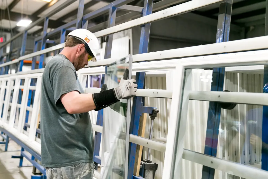A Clayton home builder in construction clothes and a hard hat fits windowpanes into a white window frame.