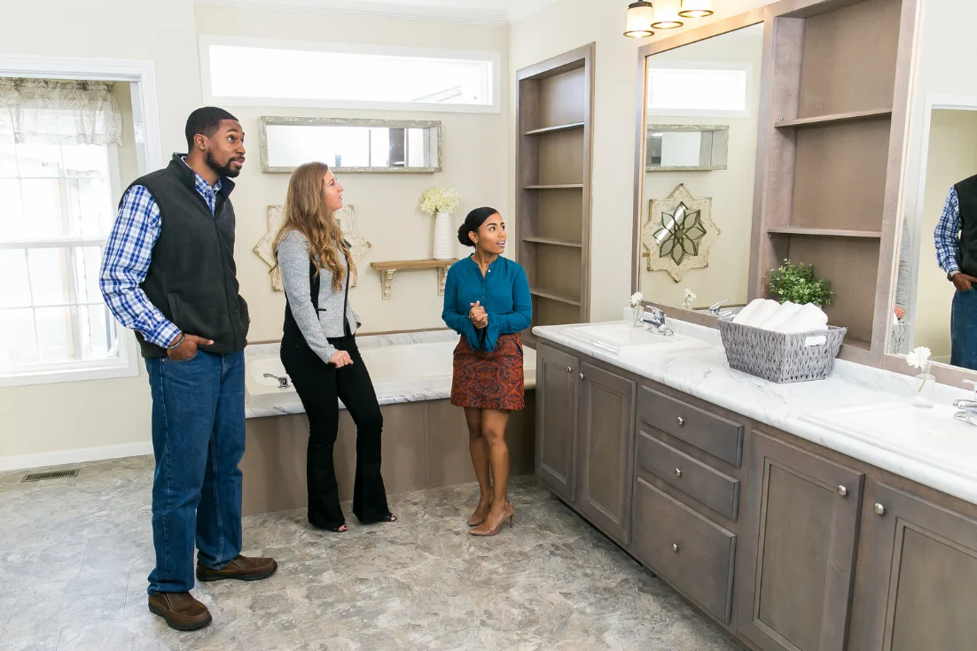 A home consultant gives a tour to a young couple of a manufactured home primary bathroom with dual sinks and built-in shelves.