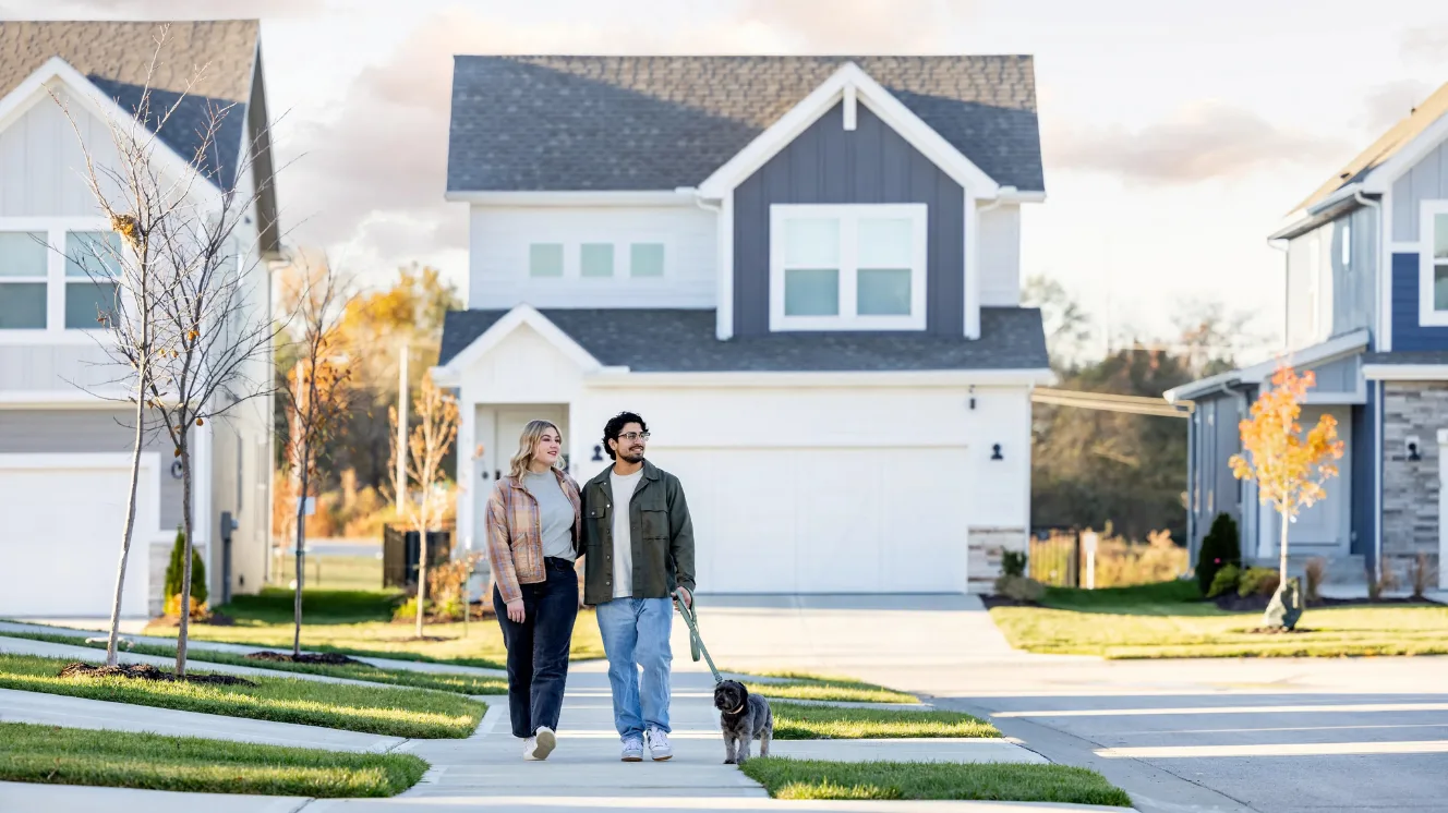A couple in jackets walks their dog down a sidewalk in a new construction neighborhood of two-story homes.