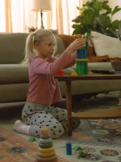 A young girl sits on the living room floor by a couch, building blocks on a wooden coffee table.