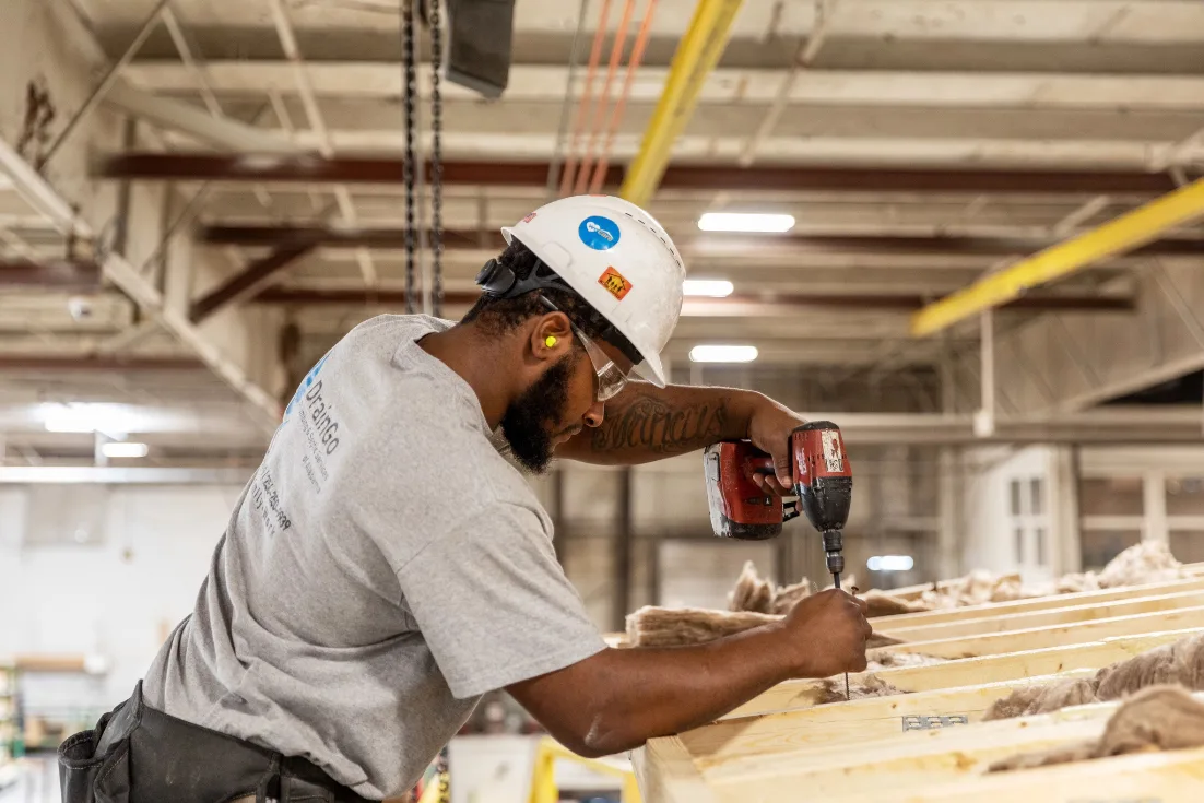 A man wearing a hard hat and safety glasses uses a drill to work on the roof of a Clayton manufactured home inside of a building facility.