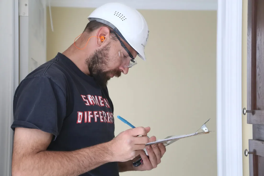 A bearded man in a dark T-shirt wears safety glasses, orange earplugs, and a white construction safety hat while writing on a clip board with a pen.