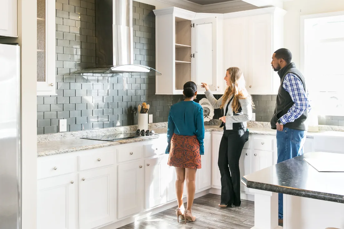A home consultant gives a tour of a large kitchen to a young couple, opening the cream colored cabinets.