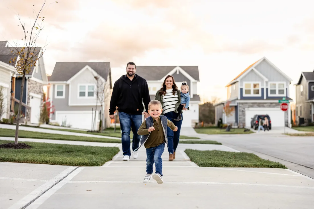 A little boy runs in front of his parents, the mom holding a toddler on her hip, on the sidewalk with two-story  homes behind them in a new construction neighborhood.