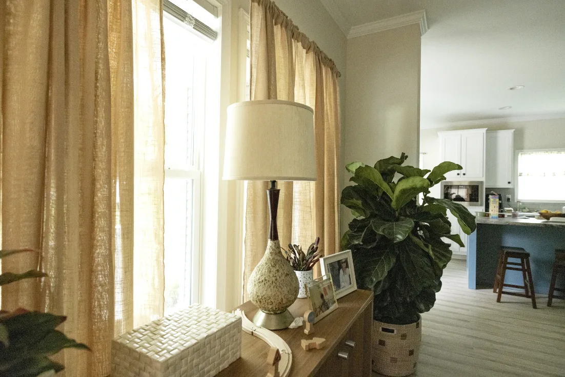 A console table with a lamp, frames, and decor is set in front of a large window with beige curtains, bright outside light shining through, in a living room with the kitchen seen in the background.