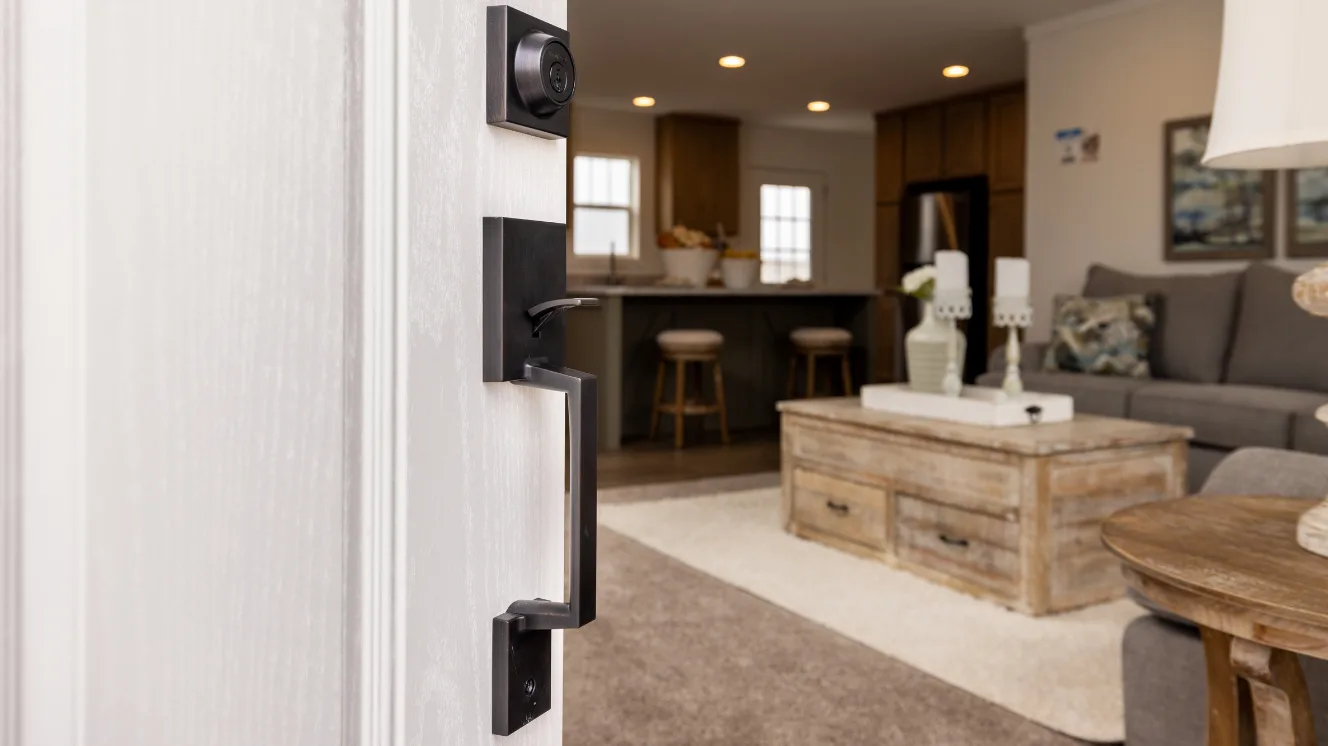 A white open front door with iron hardware opens to an open living room with a gray couch and wooden coffee table, the kitchen in the background.