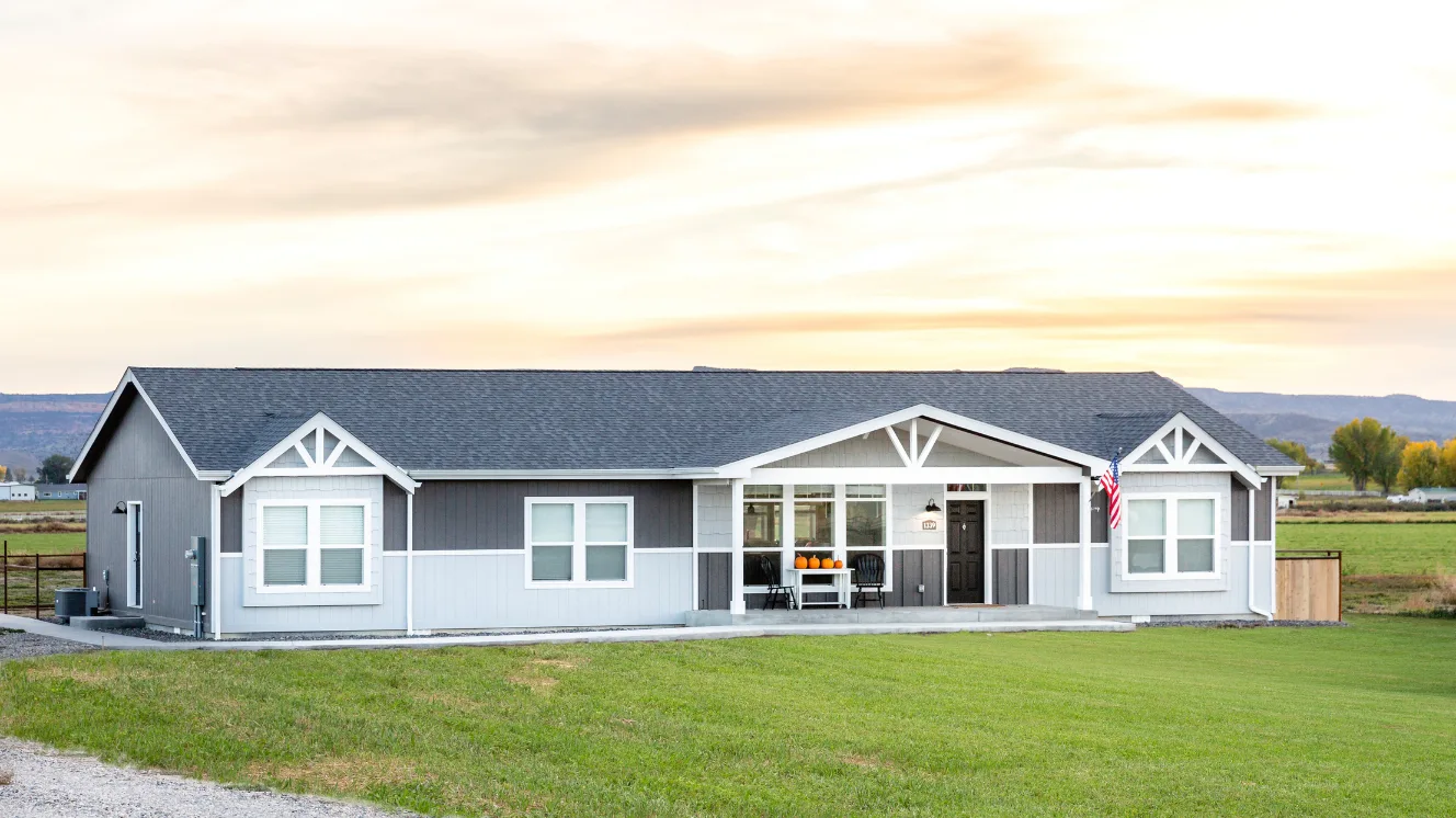 An exterior of a modern manufactured home with blue and gray siding, white dormers, and front porch on a bright green lawn.