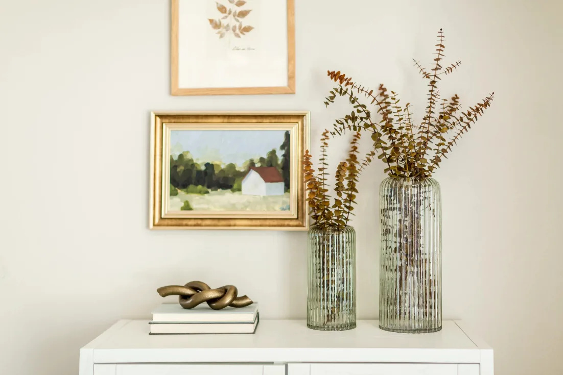 A close-up of two a book with a decorative metal knot sculpture and two glass vases sit on top of a white dresser below two small framed illustrations.