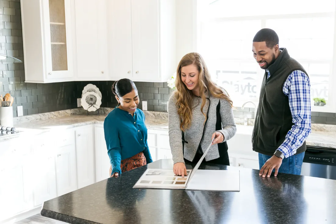 A home consultant stands between a young couple in the kitchen of a modern manufactured model and points at a picture of a countertop option in a catalog on the kitchen island.