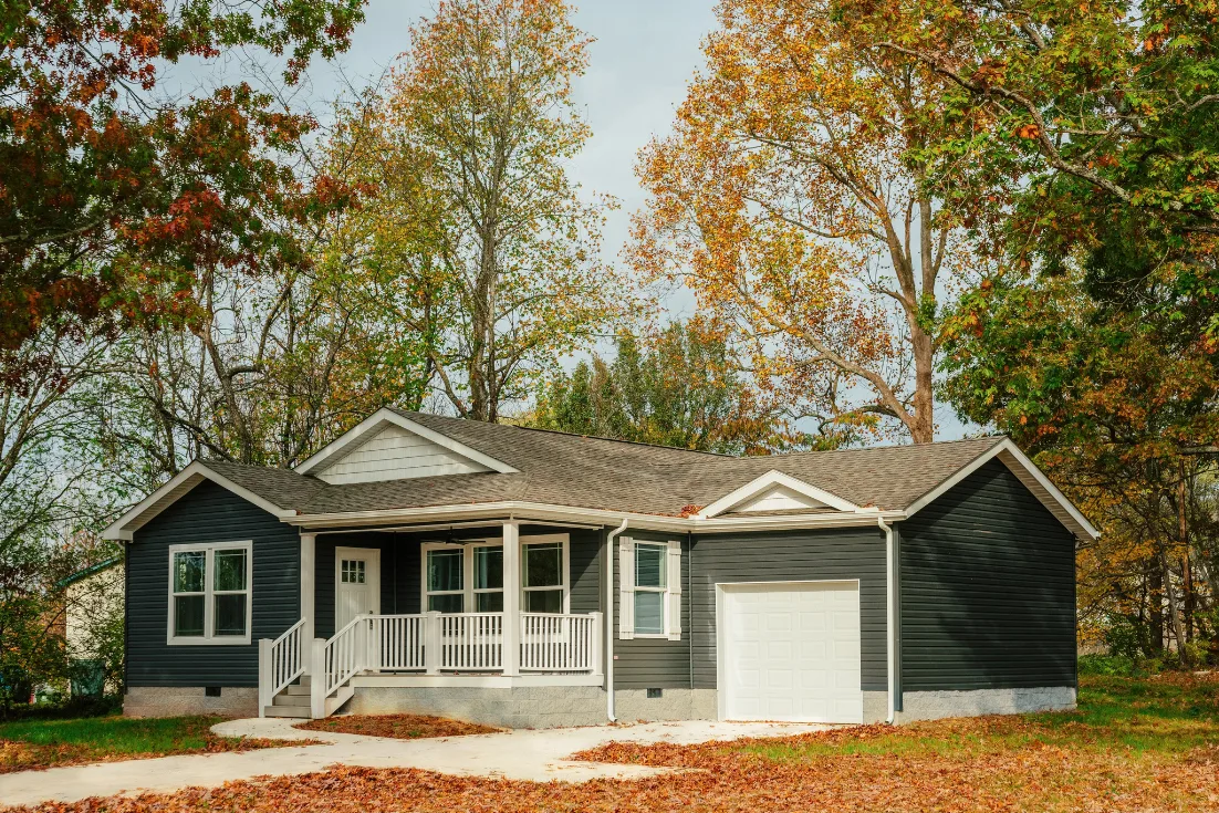 An exterior of a CrossMod® with gray siding, a front porch with white railing, and an attached garage on an autumn day surrounded by colorful trees and fallen leaves.