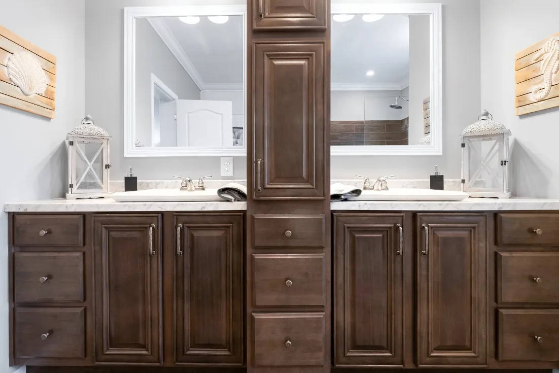 A dual vanity in a bathroom is divided by a built-in linen cabinet, dark brown cabinets, and white marble countertops.