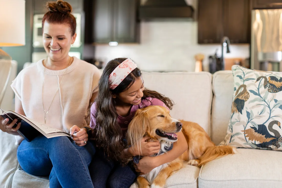 A woman on a cream sweater looks down at a magazine while sitting on a cream couch while a little girl is beside her smiling and hugging a tan, fluffy dog.