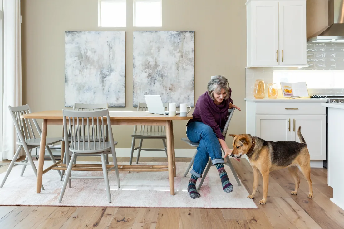 A woman sits sideways in a chair beside her laptop at the kitchen table and leans down to pet her brown and black dog.
