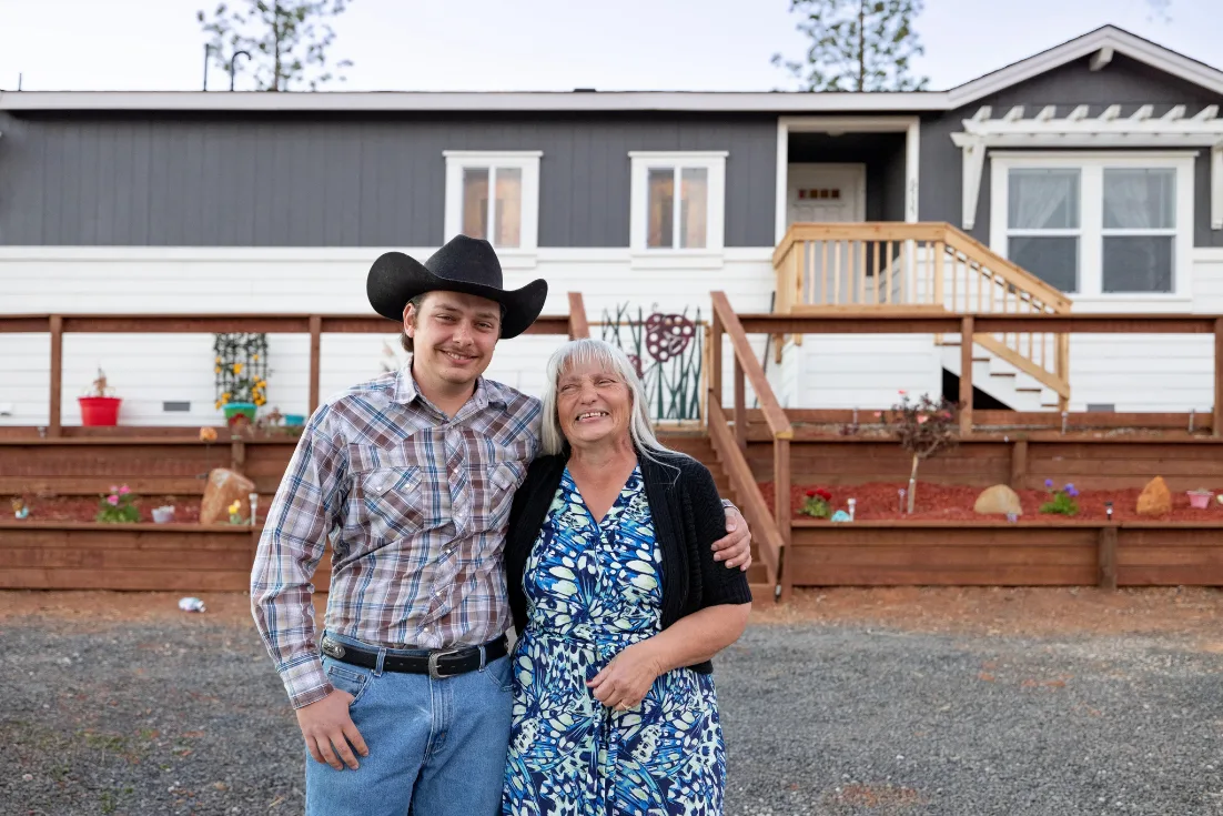 Shirley Birdsong, in a blue  print dress, and her son, Salvador, wearing a plaid shirt and cowboy hat,  pose in front of their gray and white modern manufactured home.