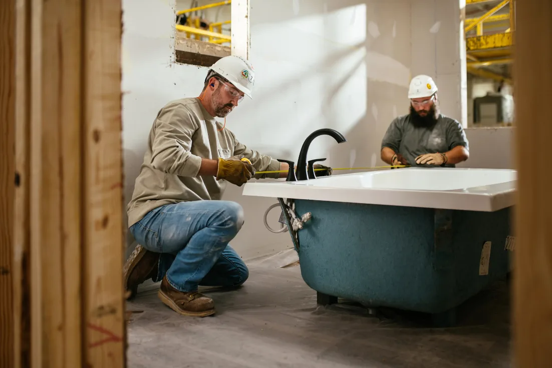 Two facility workers in hard hats install a bathtub in an unfinished new construction home.