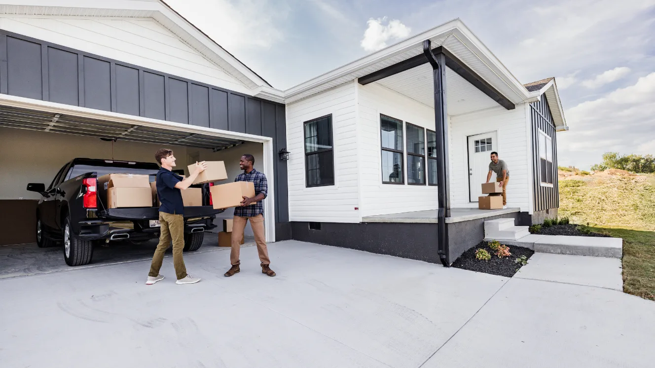 Two men pick up moving boxes from a black truck parked in an open garage attached to a white and gray CrossMod® home, while another man picks up boxes at the front door on the side of the front porch.