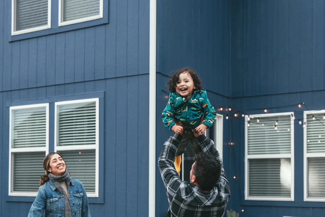 A dad holds up his laughing toddler son while the mother in a jean jacket watches and smiles.