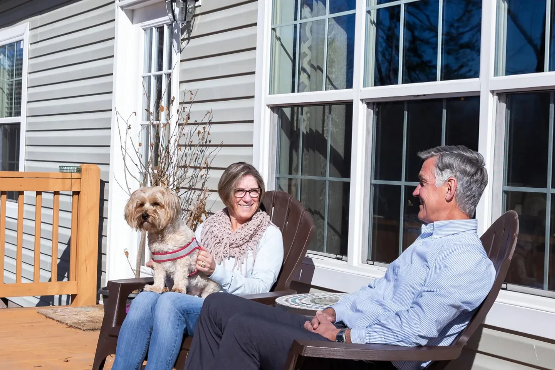 Rob and Katie Allyn sit in brown Adirondack chairs with their small tan dog in Katie's lap while on the front porch of their modern manufactured home.