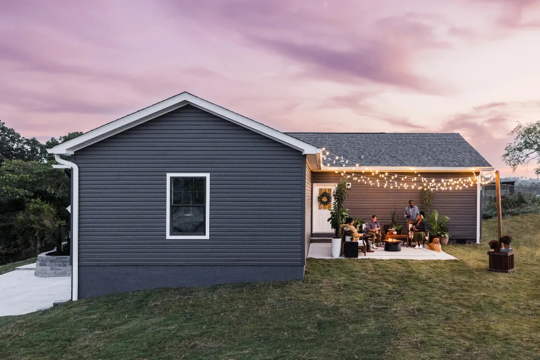 The back of a new construction home showing a patio party at dusk with friends around a fire pit under string lights.