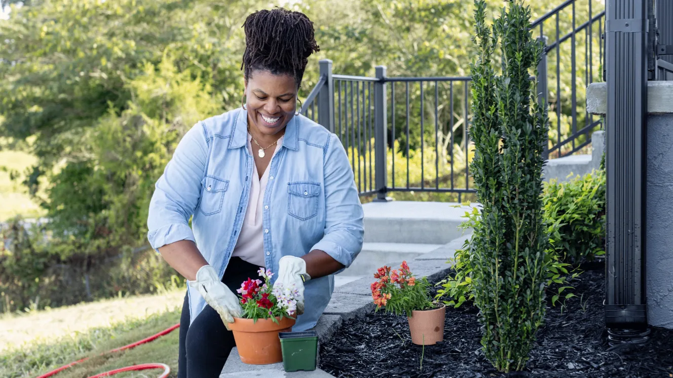 A woman in a denim shirt and gardening gloves adjusts flowers in a pot beside her raised garden bed.