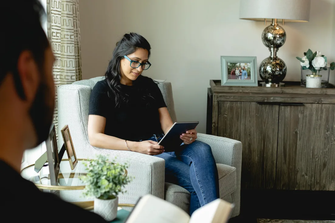 A woman in a black shirt and glasses sits in a beige armchair looking down at a tablet in her hand.