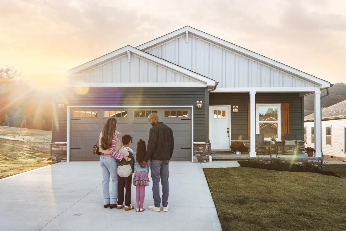A family of four stands in their driveway at sunset and faces their home with a gray garage door, a front porch, blue siding, and a white front door.