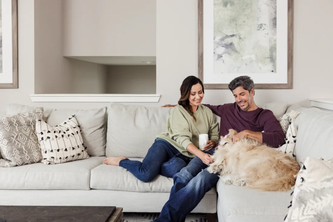 Husband and wife sit inside their new construction living room with their dog.