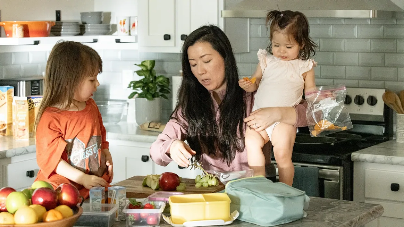 A mother holds a toddler at a kitchen island while preparing fruit, her other young daughter watching.