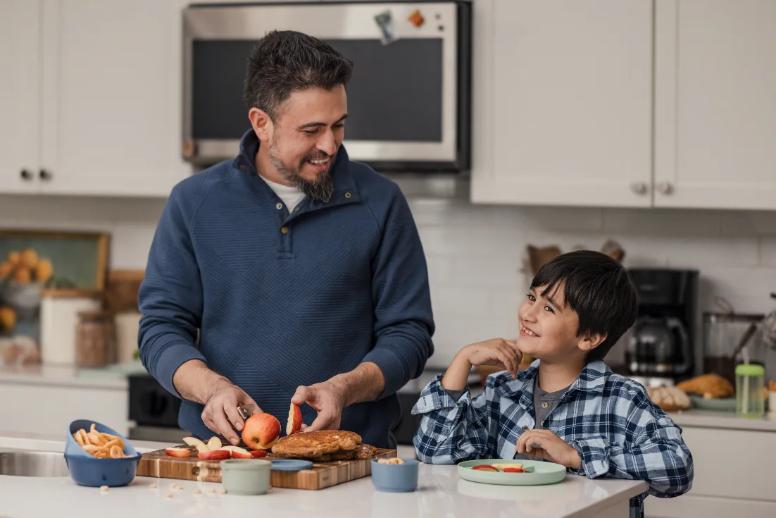 A dad in a blue pullover sweater smiles at his son in a blue flannel shirt beside him as he prepares sliced apples at the kitchen island.