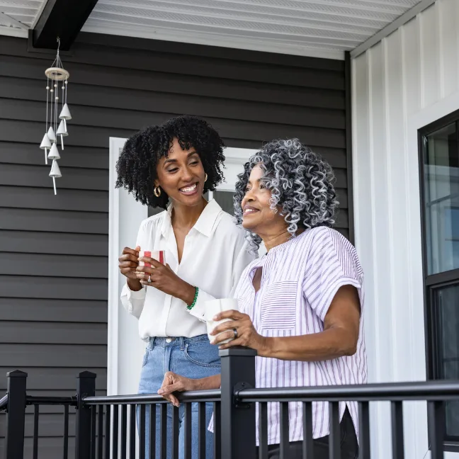 A daughter and her older mom stand on the front porch smiling and holding coffee cups.