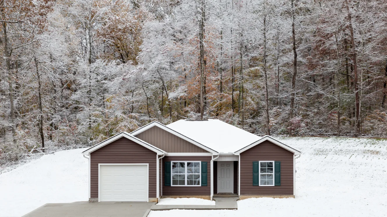 An exterior of a CrossMod® home with brown siding and an attached garage on a snowy lawn with winter-bare trees in the background.