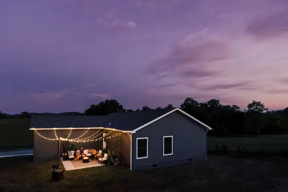 The back exterior of a CrossMod® home under an evening sky with a group sitting around a firepit on the patio with string lights hanging above them.