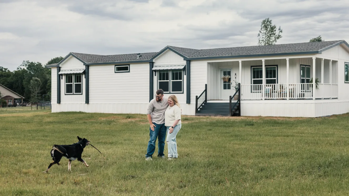 Koven and Josh Bruce standing outside their manufactured home while their dog Lina brings them a stick.