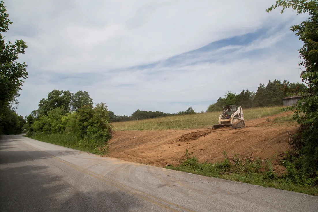 A skid steer moves dirt to clear land off a paved road.